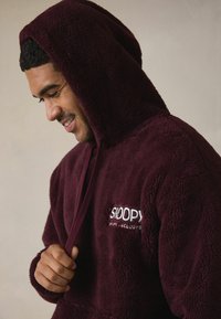 Bordūras krāsas kapuci vilnas džemperis ar izšūtu logotipu, mīkstu tekstūru un auklu. Ir brīvs siluets un garas piedurknes.