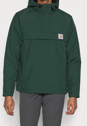 Mężczyzna ubrany w ciemnozieloną kurtkę Carhartt z kapturem i przednią kieszenią oraz szare spodnie, stojący na jednolitym jasnym tle.
