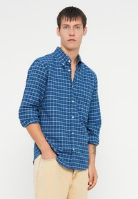 Camisa de cuadros azules de botones en algodón, con mangas largas, cuello con solapa, bolsillo frontal y dobladillo redondeado. Combinada con pantalones beige.