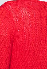 Röd stickad tröja med ett strukturerat, kabelmönster. Tyget har ett mjukt utseende med en något öppen v äv, vilket betonar värme.