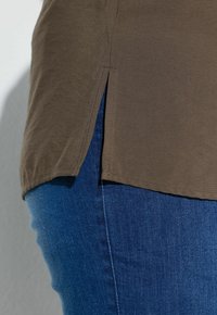 Ourlet de chemise marron avec fente latérale portée sur un jean en denim bleu au niveau de la hanche et du haut de la cuisse d'une personne.