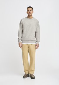 Grå sweatshirt med ribbede mansjetter og fald, kombinert med beige bukser og brune sneakers. Modellen står foran en ensfarget bakgrunn.