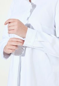Camicia bianca con bottoni, caratterizzata da un colletto e polsini arricciati, realizzata in un tessuto liscio. I polsini hanno una chiusura a un solo bottone e un design minimale.