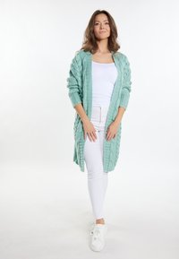 Mintgrön stickad cardigan med ett texturerat mönster, öppen front och långa ärmar, bärs över en vit tanktopp och vita byxor.