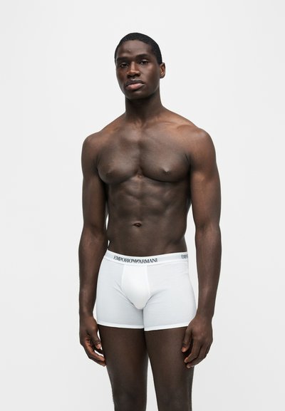 Λεπτός μαύρος άνδρας όρθιος, φοράει λευκά boxer briefs Emporio Armani, μπροστά σε απλό ανοιχτό γκρι φόντο.