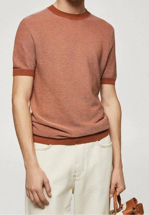 Mężczyzna w rdzawym, krótkim swetrze z drobnymi paskami i kremowych spodniach, trzymający brązową skórzaną torbę.