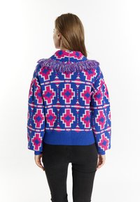 Färgglad stickad tröja med blå, rosa och vita geometriska mönster, med en fransdetalj vid kragen. Ribbat avslut och ärmslut.