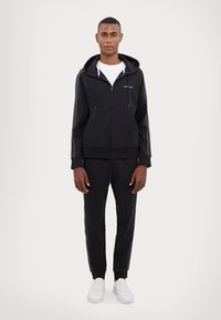 Black zip-up hoodie in cotton blend with white side stripes, paired with matching joggers. Features a kangaroo pocket and ribbed cuffs. White sneakers.