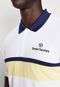 Fehér polo póló sötétkék gallérral. Sárga vízszintes csíkkal és fekete "Sergio Tacchini" logóval rendelkezik a bal oldalon. Texturált anyag.