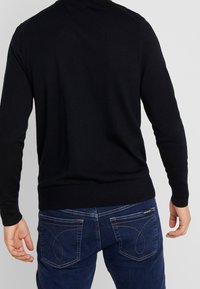 Maglione nero lavorato a maglia con maniche lunghe, design aderente, polsini e orlo a costine. Indossato sopra jeans blu scuro con cuciture in contrasto.