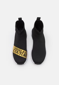 Svarta stickade högst upp sneakers med en guld "VERSACE JEANS COUTURE" logoband och vita sulor. Har en dragflik på hälen.