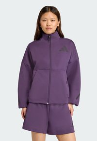 Fialová zip-up bunda s ladícími šortkami, vyrobená z texturované tkaniny; má vysoký límec a čierne akcenty, včetně loga na hrudi.