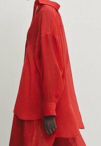 Chemise rouge oversize avec col, en tissu léger texturé. Manches longues avec poignets boutonnés et ourlet arrondi.