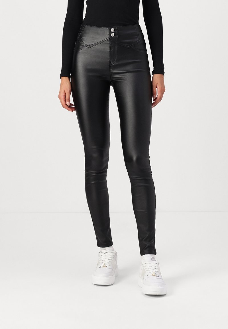 Pantalon slim noir effet cuir avec double bouton et détail zip, porté avec des baskets blanches et un haut noir à manches longues.