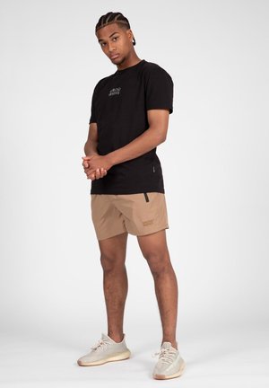 Zwarte katoenen T-shirt met witte tekst, gecombineerd met tan shorts met een zijzak en tan sneakers van gestructureerde stof.
