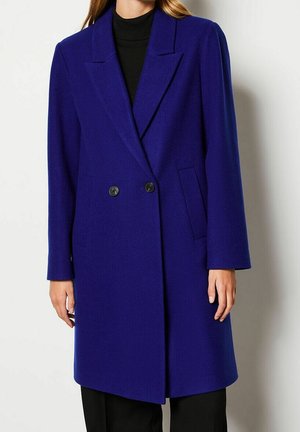 Femme portant un manteau bleu roi croisé longueur genou avec des boutons noirs sur un col roulé noir et un pantalon noir.