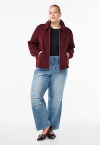 Veste en polaire bordeaux avec une fermeture éclair frontale et un col, assortie à un pantalon en denim bleu clair à jambes larges avec des coutures en panneau et des mocassins marron foncé.