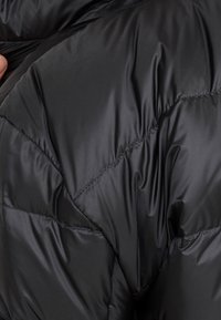 Nahaufnahme einer Hand, die eine schwarze gesteppte Daunenjacke hält und die glänzende, genähte Stoffstruktur zeigt.