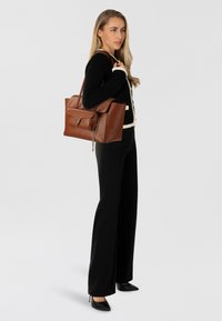 Bruine leren handtas met een gestructureerde vorm, voorzien van een flapsluiting en een kettingaccent. Het model draagt een zwarte gebreide trui en zwarte broek.