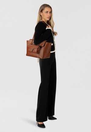 Borsa a mano in pelle marrone con una forma strutturata, dotata di chiusura a patta e dettaglio a catena. Il modello indossa un maglione nero lavorato a maglia e pantaloni neri.