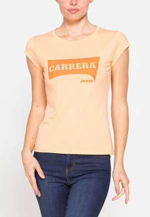 Donna che indossa una T-shirt aderente color pesca con il logo "CARRERA Jeans" e jeans blu scuro, in piedi davanti a uno sfondo chiaro e uniforme.
