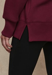 Bordeauxfarbener Pullover mit gerippten Manschetten und seitlichen Schlitzen, kombiniert mit schwarzen Leggings. Der Stoff wirkt weich und strukturiert.