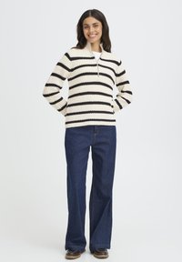 Pletený sveter s krátkym zipsom, s čierno-krémovými horizontálnymi pruhmi, skombinovaný s modrými riflovými rozšírenými nohavicami a hnedou obuvou.