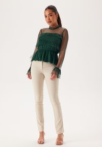 Groene blouse met gestructureerde doorschijnende mouwen, gerimpeld ontwerp en een getailleerde taille, gecombineerd met crèmekleurige slim-fit broeken en nude sandalen.