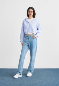 Φως μωβ cropped φούτερ με φερμουάρ μπροστά και τσέπες, σε συνδυασμό με μπλε παντελόνι φαρδύ με κάθετες ρίγες και λευκά αθλητικά παπούτσια.