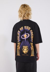 Black oversized T-shirt featuring a colorful graphic design with pillars and classical imagery, "Legacy of the Cosmos" text, and vibrant accents.