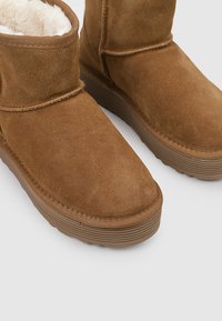 Par de botas de invierno de ante marrón con suelas de goma gruesas y suave forro de lana blanca, mostradas sobre un fondo gris.