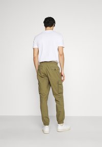 Pantalon cargo kaki avec poignets élastiques et multiples poches, associé à un t-shirt blanc à col rond et des baskets blanches, vu de dos.