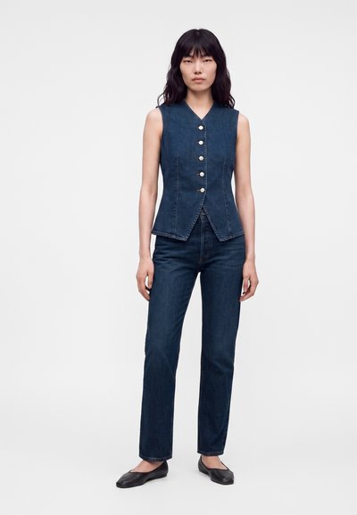 Femme debout portant un gilet en jean bleu foncé sans manches avec des boutons argentés, un jean bleu foncé assorti et des chaussures plates noires sur fond blanc.