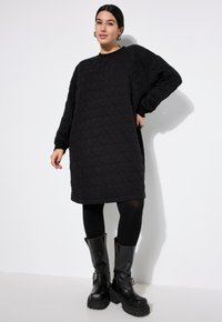Vrouw in een zwart gewatteerd sweatshirtjurk met hartpatroon, zwarte leggings en zwarte stevige gespschoenen tegen een witte achtergrond.