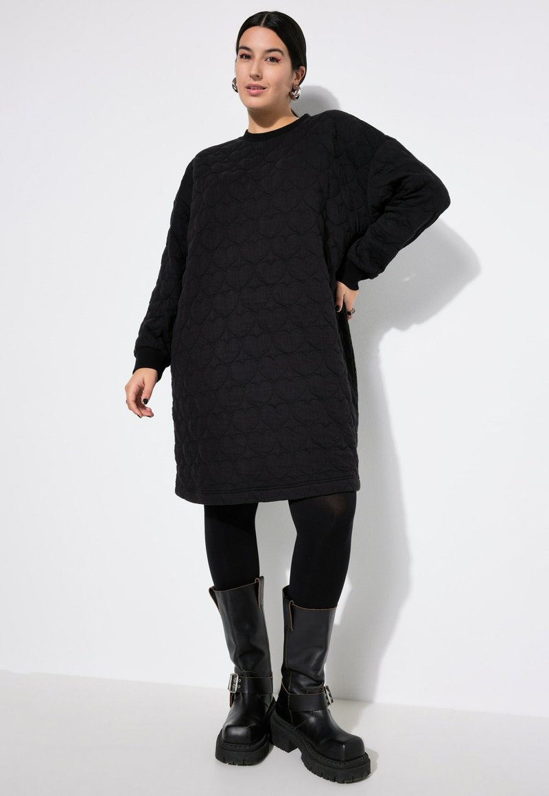 Vrouw in een zwart gewatteerd sweatshirtjurk met hartpatroon, zwarte leggings en zwarte stevige gespschoenen tegen een witte achtergrond.