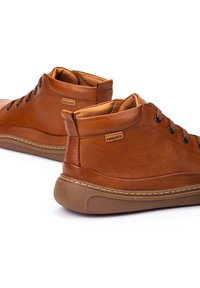 Par de zapatos casuales de cuero marrón de media altura con cuellos acolchados, diseño con cordones y etiquetas de marca en el lateral y en las suelas del talón.