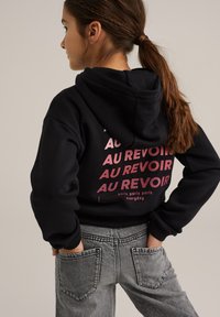 Črna kapuča s puloverjem z roza grafičnim besedilom "AU REVOIR" v različnih velikostih; v kombinaciji z sivo jeans hlačami, ki imajo zadnje žepe.