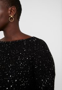 Maglione nero con paillettes e scollo a barca; grandi orecchini rotondi dorati che valorizzano il design. Attenzione alla texture e alla brillantezza dei materiali.