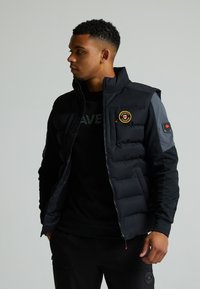 Gilet imbottito nero con colletto alto, motivo trapuntato sul corpo e maniche con accentuazione grigia. Presenta un badge con logo sul petto e polsini a coste.