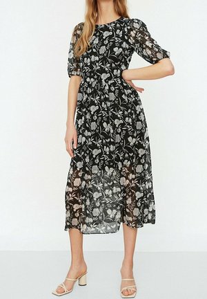 Frau trägt ein schwarzes Midikleid mit weißen Blumenmustern und transparenten Puffärmeln, kombiniert mit beigen offenen Sandalen mit Blockabsatz.