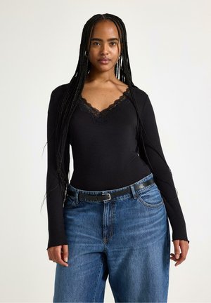 Ung kvinde med langt flet hår iført en sort top med blonder og blå jeans med sort bælte, stående foran en ensfarvet baggrund.