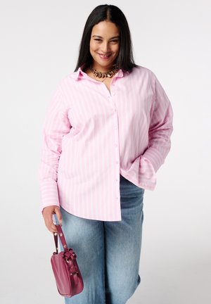 Vrouw die glimlacht en een roze gestreept overhemd met knopen draagt, lichtblauwe jeans, een gouden ketting en een kleine paarse handtas naast zich houdt.