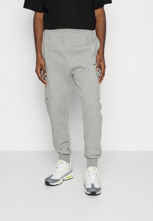 Mann trägt hellgraue Cargo-Jogginghosen mit Seitentaschen und weiße-graue Sneaker mit neongrünen Akzenten, steht vor weißem Hintergrund.