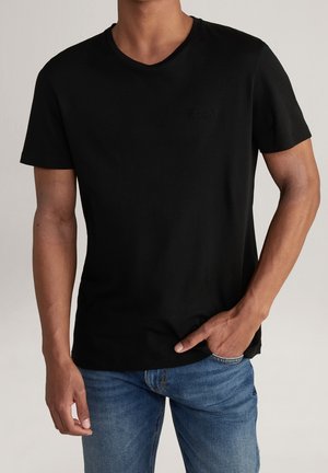 Homme portant un t-shirt noir uni à manches courtes et un jean bleu, une main dans la poche, sur un fond neutre.