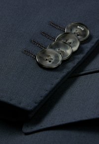 Tessuto della giacca blu navy con superficie liscia e cuciture visibili. Quattro bottoni scuri, contrassegnati "BOSS," disposti in modo ordinato lungo l'apertura.