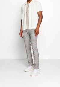 Hedvábná košile s krátkým rukávem slonovinové barvy přes béžové tričko, spárovaná se šedými kostkovanými slim-fit kalhotami a bílými teniskami.