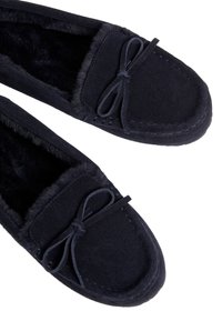 Mocasin de ante azul marino con forro de piel sintética, acento de lazo y detalle de costura. Textura suave con diseño de puntera redondeada.