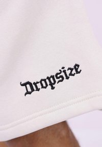 Hvid bomuldsstof med sort broderet tekst "Dropsize" ved kanten. Teksturen virker glat med en let glans.