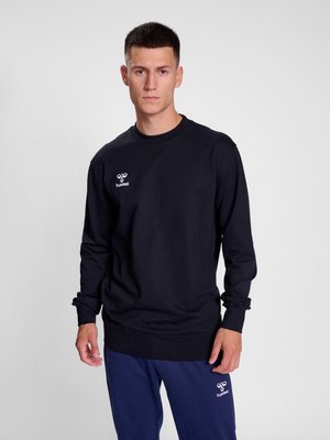 Junger Mann, der einen schwarzen hummel Langarm-Sweatshirt und marineblaue hummel Hosen trägt, steht vor einem schlichten hellgrauen Hintergrund.