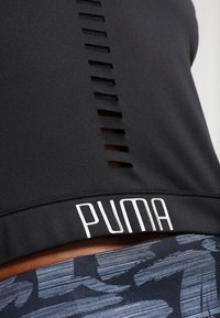 Schwarzes Sportoberteil mit Ausschnitt auf dem Rücken und weißem "PUMA"-Logo am Saum. Getragen mit gemusterten, dunkelblauen Hosen mit abstrakten Strichen.
